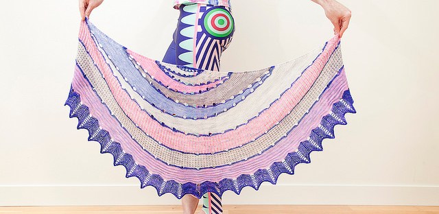 Stephen West, wearing multicoloured top and leggings and holding knitted Exploration Station shawl in pastel pinks, purples and yellows