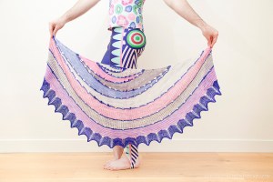 Stephen West, wearing multicoloured top and leggings and holding knitted Exploration Station shawl in pastel pinks, purples and yellows