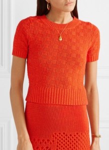 Woman wearing bright orange, short, fitted, knitted top with all-over, chequerboard texture and deep ribbing at hem.
