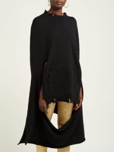 Woman wearing knitted, black sweater or poncho with roughly finished edges, and large cut outs that give distressed finish.