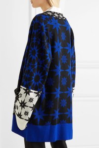 Model wearing long, knitted cardigan with rich intarsia colour-work star designs in shades of blue and with contrast white sleeve detail and blue hem.