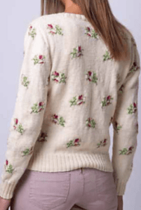 Woman wearing knitted white, fitted cardigan with floral intarsia motifs in red and green repeated across the fabric.