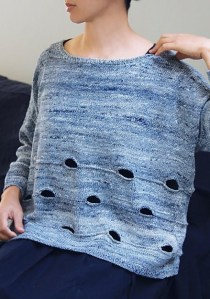 Woman wearing oversized, knitted sweater with large eyelet rows across bottom half of body and with drop sleeves and asymmetric hem.