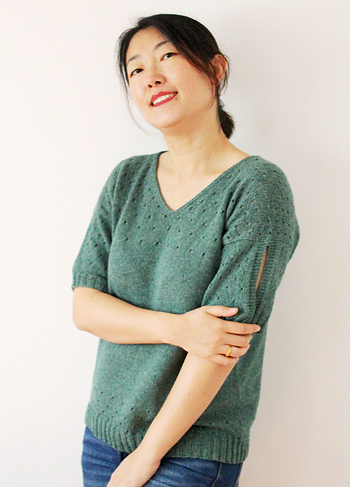 Woman wearing sage green, knitted top made out of wool/cotton blend; the top has a v-neck, and eyelet lace detail at the top section and hem, plus cut-out sleeve detail.
