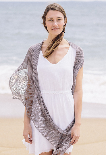 Woman standing on a beach. She is dressed in a white dress and wearing a knitted, grey shawl worked in brioche stitch and with a wide lace border.