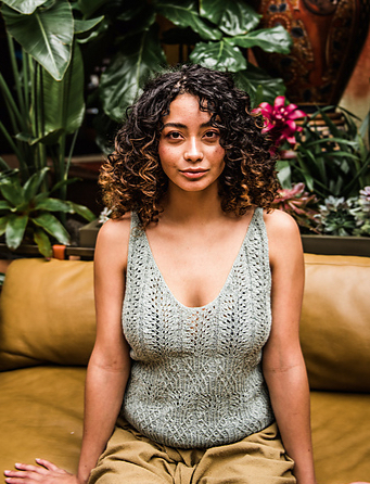Woman sitting on ochre sofa in front of green plants, wearing grey, knitted lace tank top with v-neck and ochre trousers.