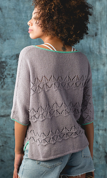 Woman wearing shorts and a lavender grey knitted sweater made from cotton/linen blend and with repeated lace motif stripe, and contrasting turquoise green edging at neck and sleeve cuffs.